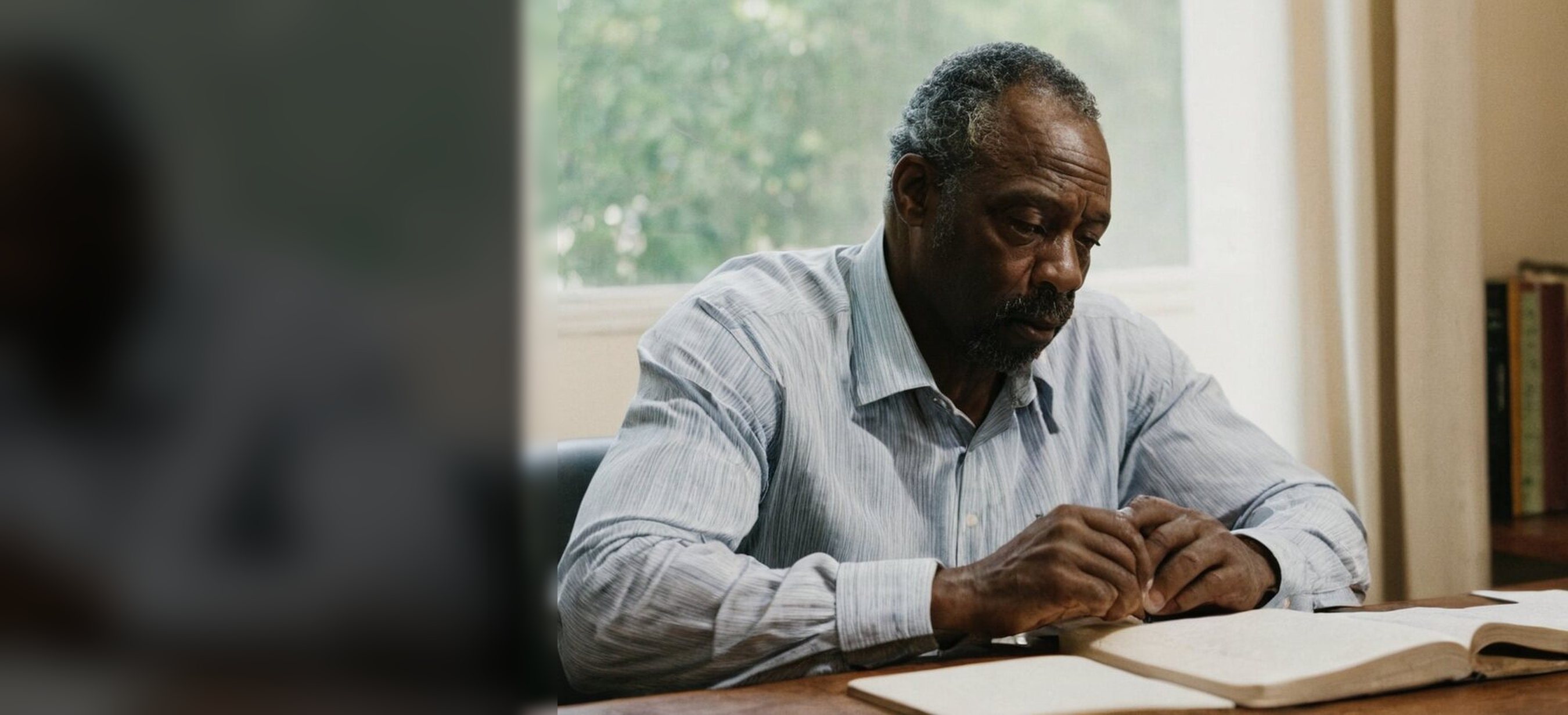 A man in his sixties reading an open Bible at a desk by a window